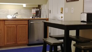 A kitchen with wooden cabinets, a sink, and a coffee maker. There is a dining table with two stools and a framed picture on the wall.