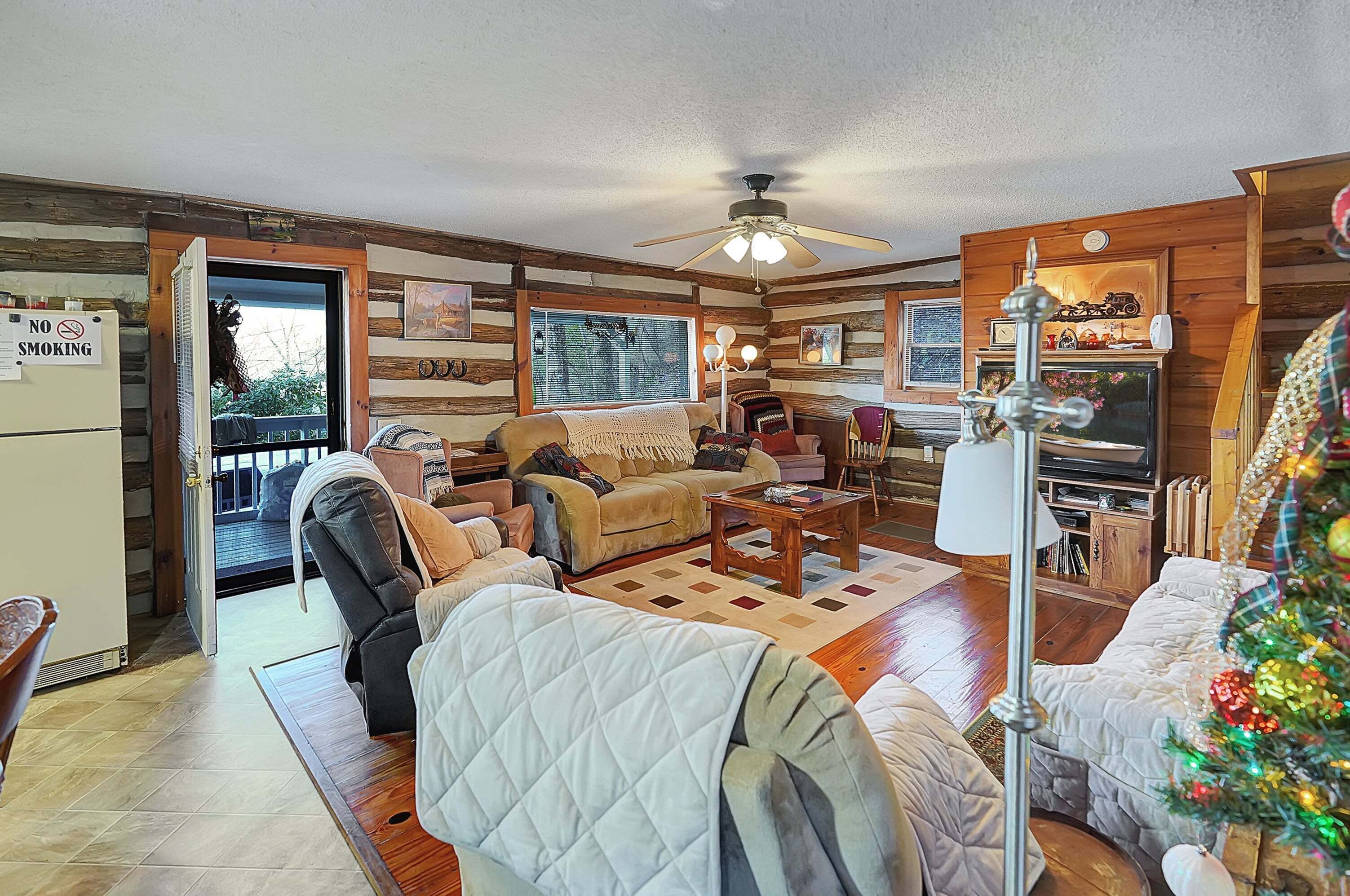 A cozy cabin interior with a fireplace, a ceiling fan, and a Christmas tree.