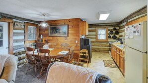 Interior acolhedor de uma cabana, com uma mesa de jantar de madeira, cadeiras, lareira e cozinha.