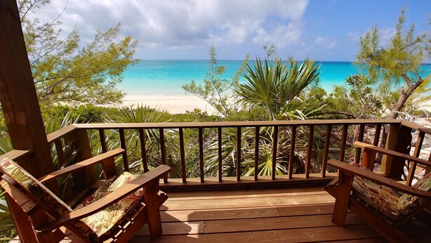 Una terraza de madera con dos sillas y un banco, con vista a una playa de aguas turquesas cristalinas y palmeras.