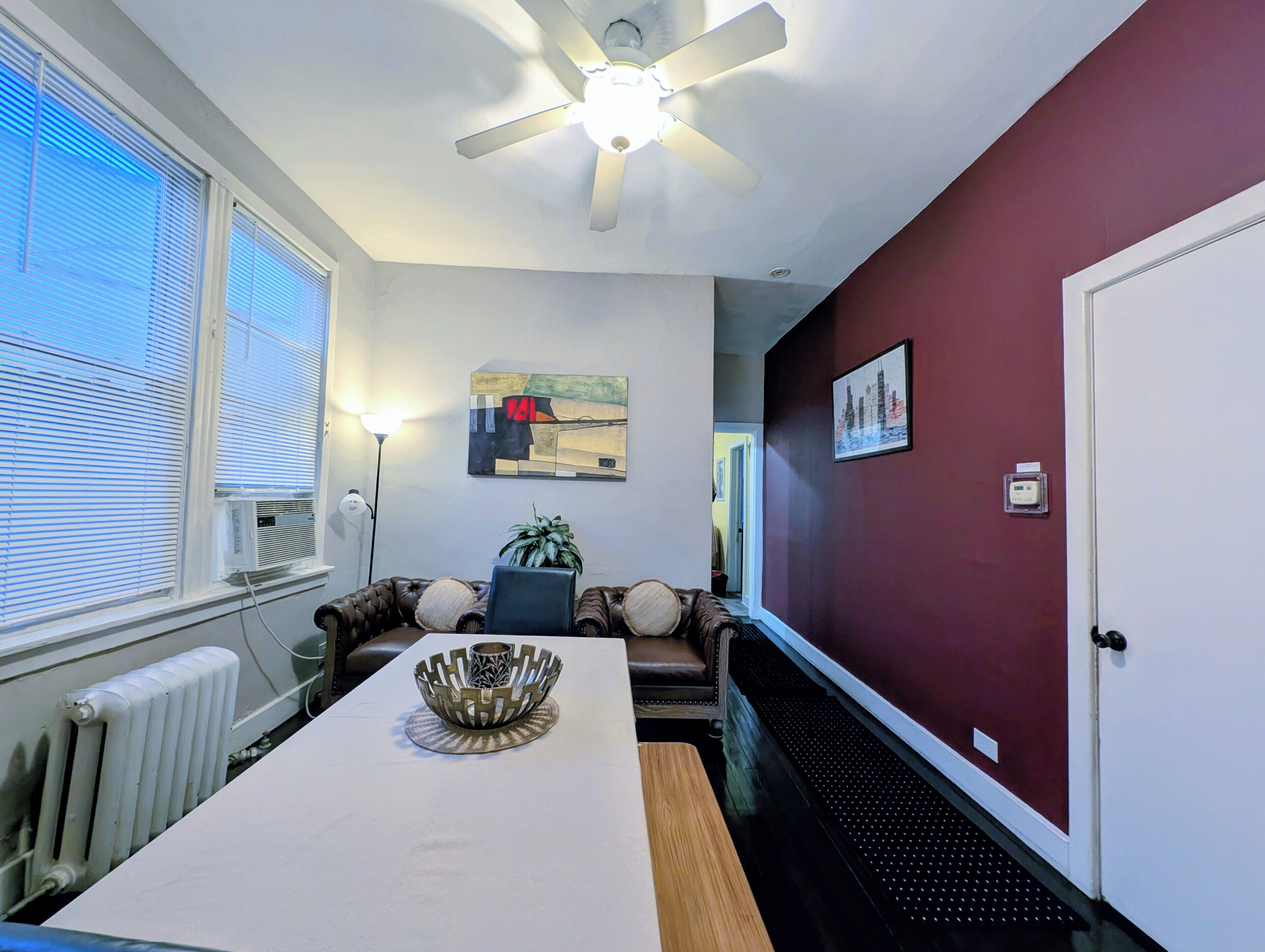 Une pièce avec un ventilateur de plafond blanc, une table sur laquelle est posé un vase décoratif, un canapé en cuir marron et un mur d’accent rouge.