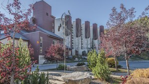 A modern building with a mix of red and white exterior walls, surrounded by landscaped gardens with various trees and a large rock feature. - Ski in / ski out family friendly condo w/access to pool, hot tub & game room (Winter Park)
