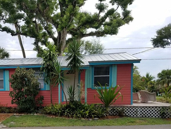 A small, single-story house with a red exterior and blue trim, surrounded by lush greenery and a white fence. - Riverfront One Bedroom Fishing Cottage w/ boat dock-Summer Specials!! (Sebastian)
