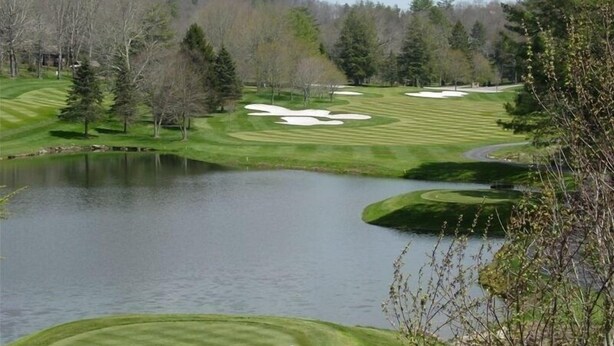 Ein Golfplatz mit Teich, Sandbunkern und einem bewaldeten Hügel im Hintergrund.