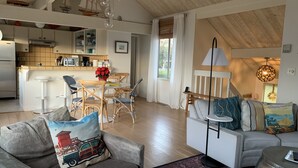 Amplia sala de estar con una mesa de comedor, sillas, un sofá y una mesa de centro. Se ve una cocina al fondo con una ventana y una lámpara colgante.