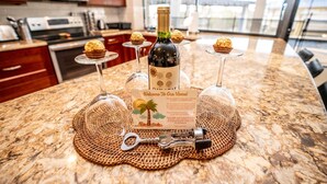 A kitchen counter with a bottle of wine, three glasses, a welcome card, and a key on a woven placemat.