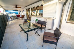 Varanda coberta com mesa e cadeiras de vidro, ventilador de teto e vista para a rua e edifícios.