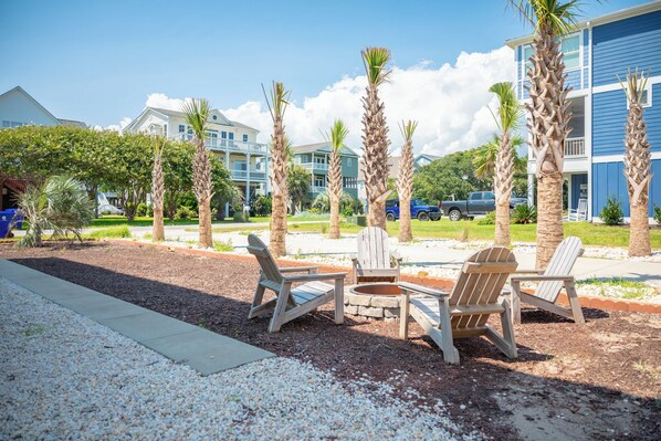 A patio area with Adirondack chairs and a fire pit, surrounded by palm trees and a gravel path. - Summer Sea: Colorful Rental with Ocean Views, Pool, Elevator (Oak Island)