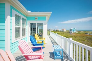 A turquoise building with a white porch, featuring colorful Adirondack chairs and a white railing overlooking a grassy area and ocean. - Summer Sea: Colorful Rental with Ocean Views, Pool, Elevator (Oak Island)