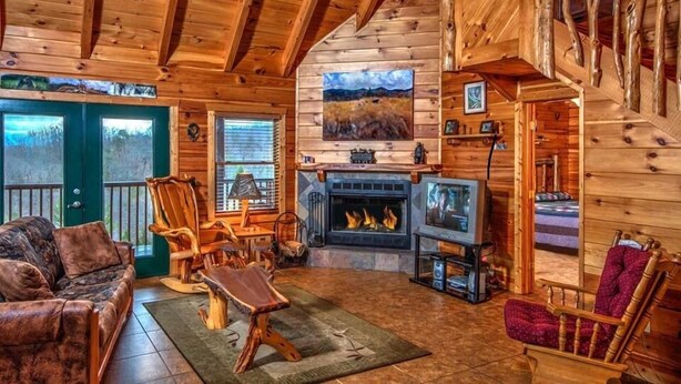 A log cabin interior with a fireplace, wooden furniture, and a large window.