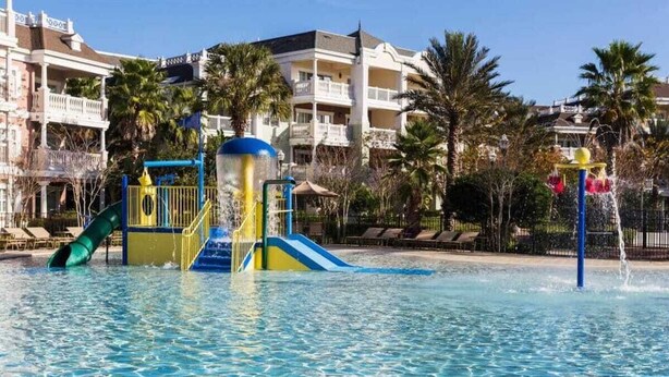 En swimmingpool med et vandlegeområde, palmer og en hotelbygning i baggrunden.