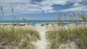 A beach with a sandy path leading to the water, surrounded by tall grasses. Beach umbrellas and tents are visible, indicating a beach setting. - Right On the Beach - Spectacular Views! (Carolina Beach)