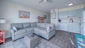 A living room with a grey sectional sofa, a small ottoman, a ceiling fan, and a kitchen area with white cabinets and a sink. - Right On the Beach - Spectacular Views! (Carolina Beach)