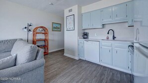 A compact living space with a kitchenette, grey sofa, wooden shelving unit, and a framed picture on the wall. - Right On the Beach - Spectacular Views! (Carolina Beach)