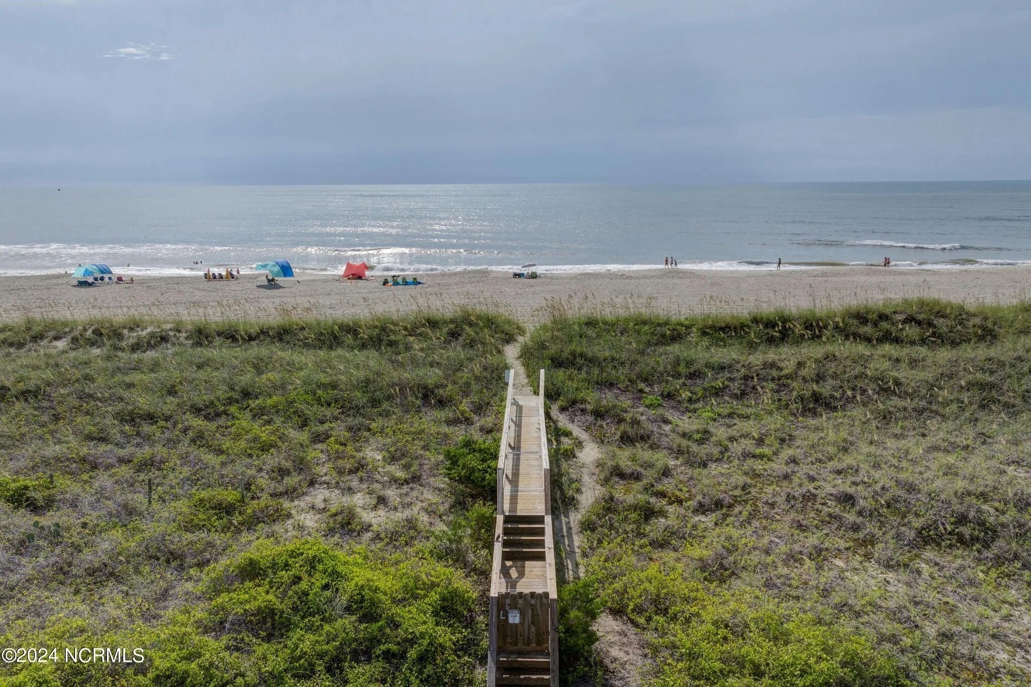 Una playa con un paseo marítimo de madera que conduce al agua, rodeada de vegetación.