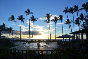 A poolside area with lounge chairs and umbrellas, surrounded by palm trees, and overlooking the ocean.