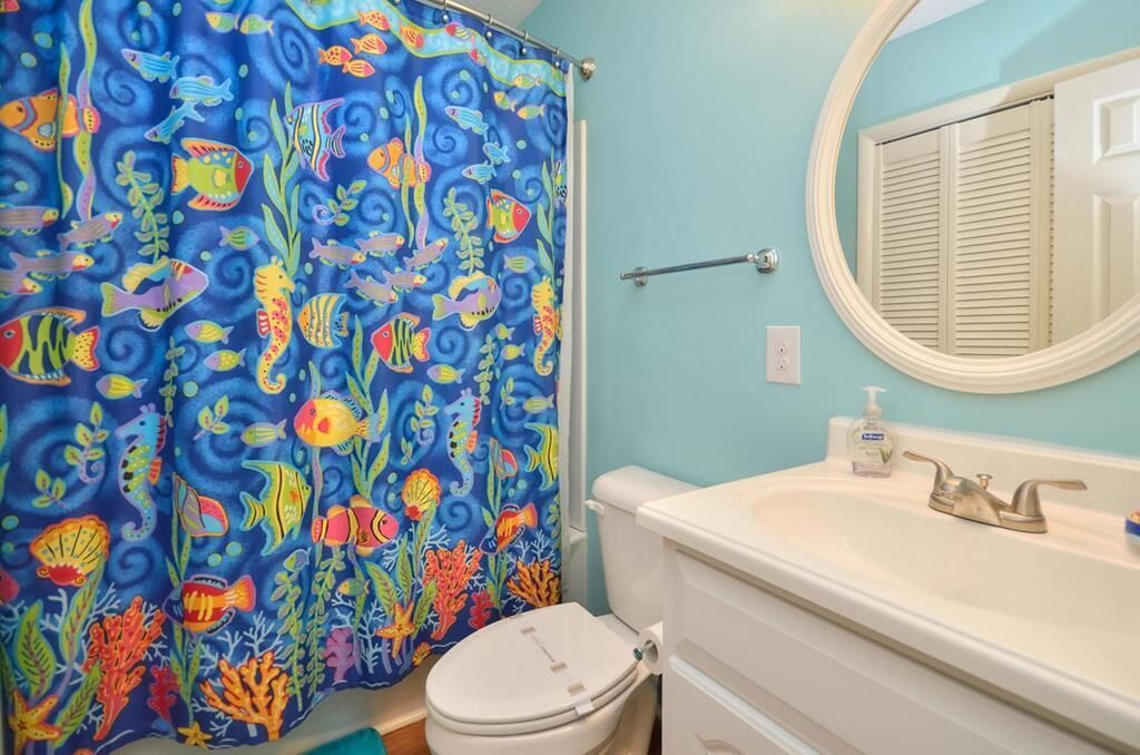 Une salle de bain avec un rideau de douche bleu orné de motifs colorés représentant des poissons et des coraux, des toilettes blanches, un miroir rond au-dessus d’un lavabo blanc muni d’un distributeur de savon, et un porte-serviettes.