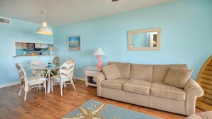 A living room with a beige sofa, a dining table with chairs, a kitchen area, and a starfish rug. - OIBV 716 - Island Dream: Oceanfront condo in Oak Island, perfect for family getaways! (Oak Island)