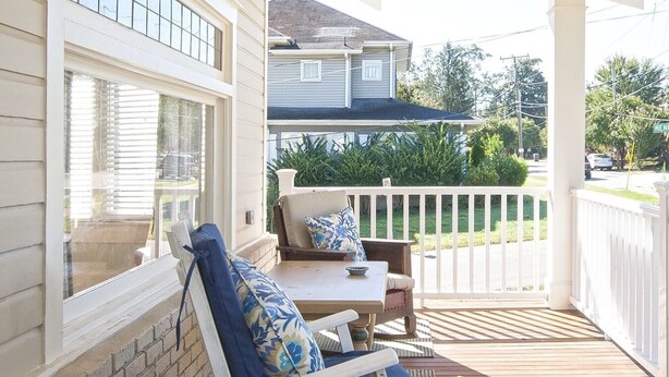 A covered porch with two rocking chairs and a table, overlooking a street and neighboring houses.