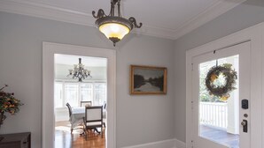 A hallway with a wooden floor, a chandelier, a dining area with a table and chairs, a framed picture on the wall, and a door with a wreath.