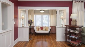A living room with a wooden floor, a white sofa with patterned cushions, a side table with a lamp, and a bookshelf.