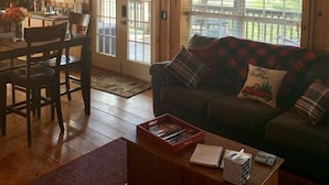 A cozy cabin interior with a wooden ceiling, a dining area with a table and chairs, a sofa with festive pillows, and a window with a red valance. - Secluded and Private Cabin with Amazing Views.  Hot tub and WiFi. (Lansing)