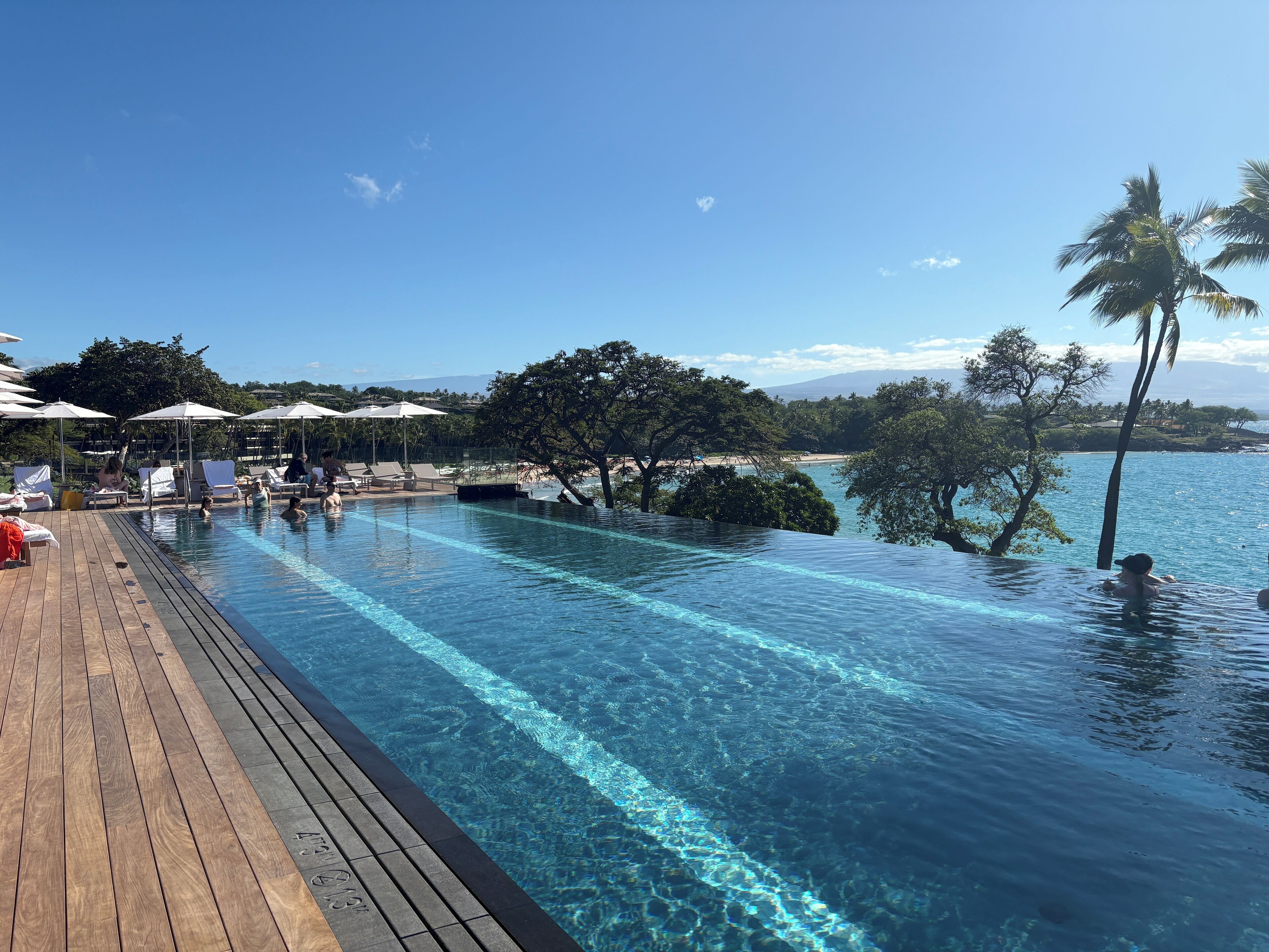 An infinity pool with clear water, wooden deck, lounge chairs, umbrellas, and a view of the beach and trees.