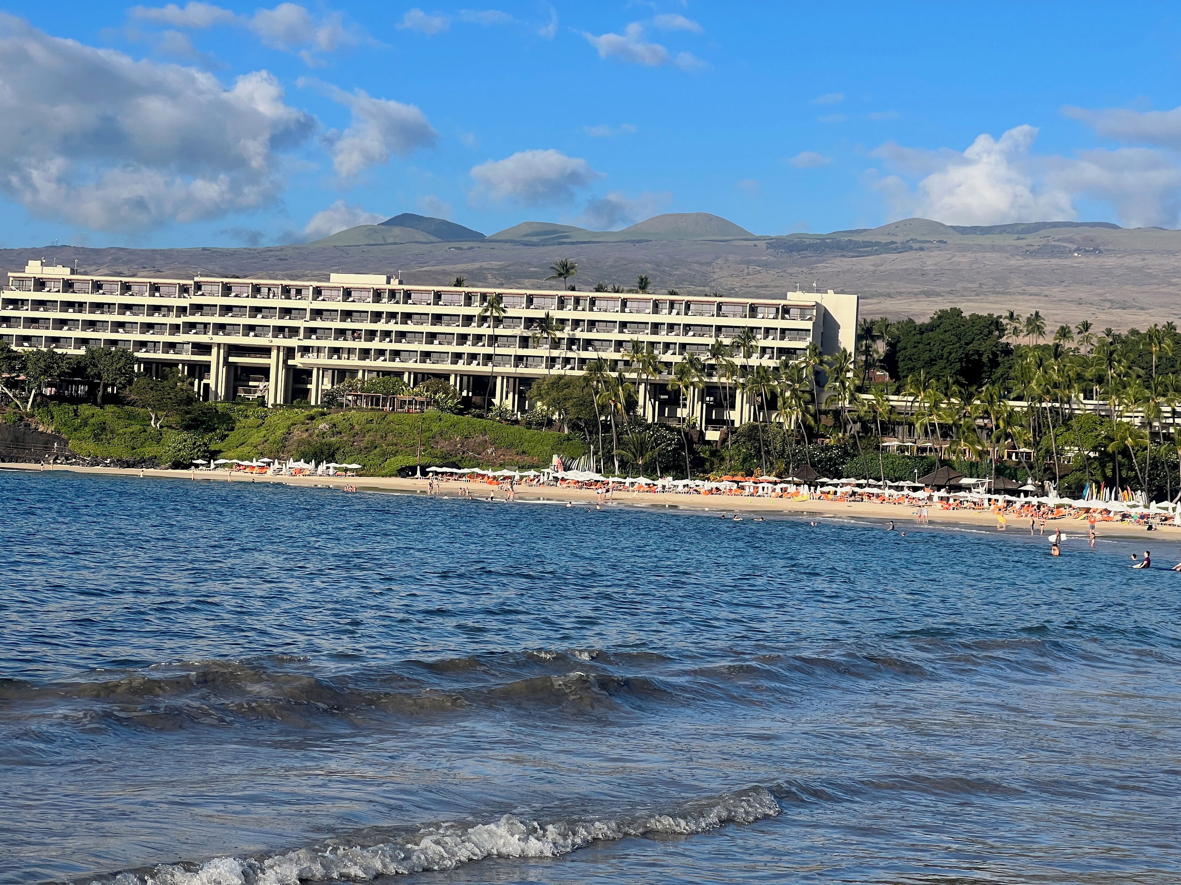 Ein Hotel direkt am Strand mit mehreren Etagen, ein Strand im Vordergrund und Berge im Hintergrund.