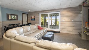 Amplia sala de estar con un sofá de cuero color beige en forma de L, un otomano negro pequeño y una mesa de comedor con sillas. Hay una puerta corrediza de vidrio que da a una terraza exterior.