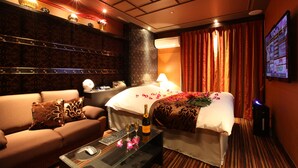Une chambre d’hôtel avec un lit, un canapé, une table basse avec une bouteille et des verres, une télévision et un mur sur lequel est fixé un écran.