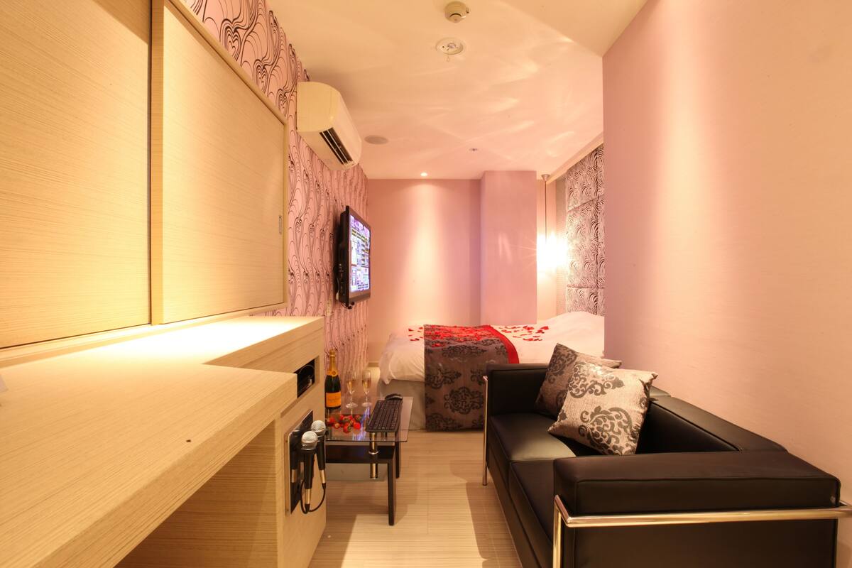 Une chambre d’hôtel compacte, dotée d’un bureau en bois, d’un canapé noir, d’un lit avec une couvre-lit à motifs et d’un téléviseur fixé au mur.
