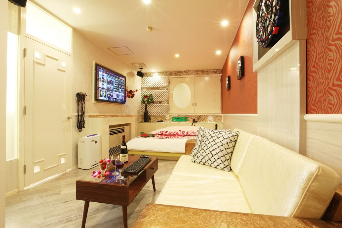A hotel room with a bed, a desk with a computer, a TV mounted on the wall, and a dartboard.