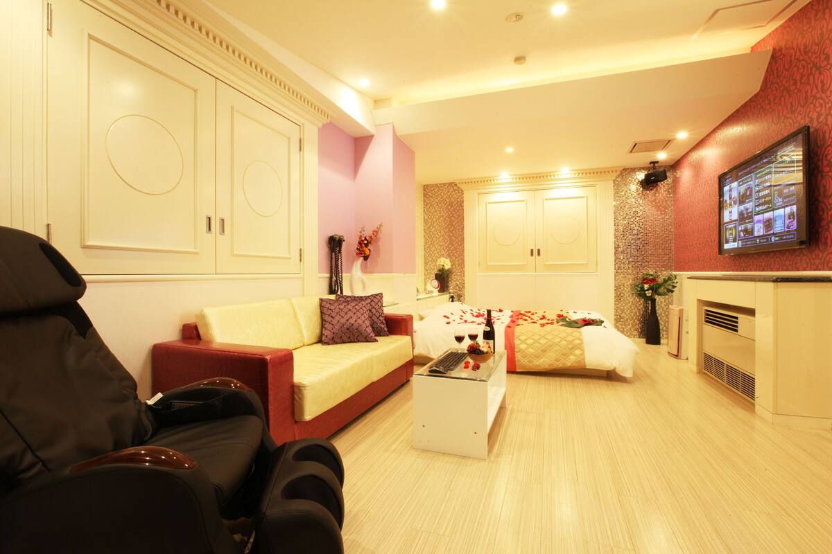 A hotel room with a sofa, a massage chair, a small table with a bottle and glasses, and a TV on the wall.