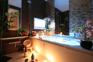 A modern bathroom with a bathtub, a sink, and a mirror. There are plants and flowers around the bathtub, and a sign with the hotel name is visible. - HOTEL GRASSINO URBAN RESORT URAWA - Adult Only (Saitama)