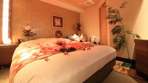 A well-lit bedroom with a large bed, floral decorations, a bamboo plant, and a painting on the wall. - HOTEL RIRAKU - Adult Only (Tokorozawa)