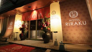 The exterior of Hotel RIRAKU with signage, a red entrance banner, and decorative vases with flowers.
