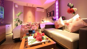 A modern living room with a sofa, a coffee table with floral arrangements, a TV, and a wall-mounted air conditioner. - HOTEL RIRAKU - Adult Only (Tokorozawa)