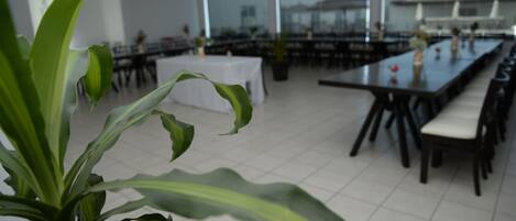 A spacious dining area with multiple tables and chairs, large windows, and a potted plant in the foreground.
