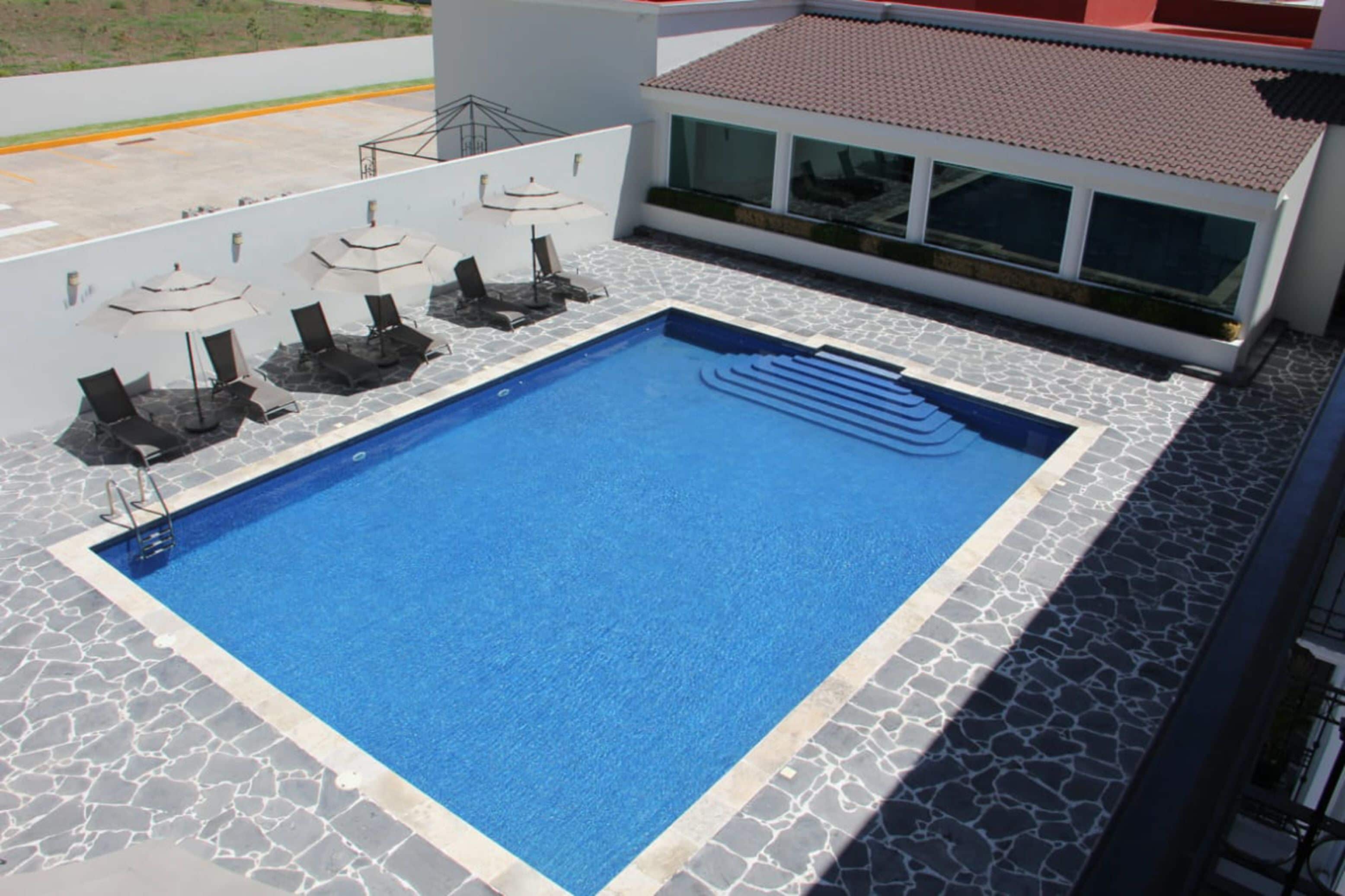 Ein rechteckiger Swimmingpool mit einer Bordüre aus Steinplatten, umgeben von Liegestühlen und Sonnenschirmen.