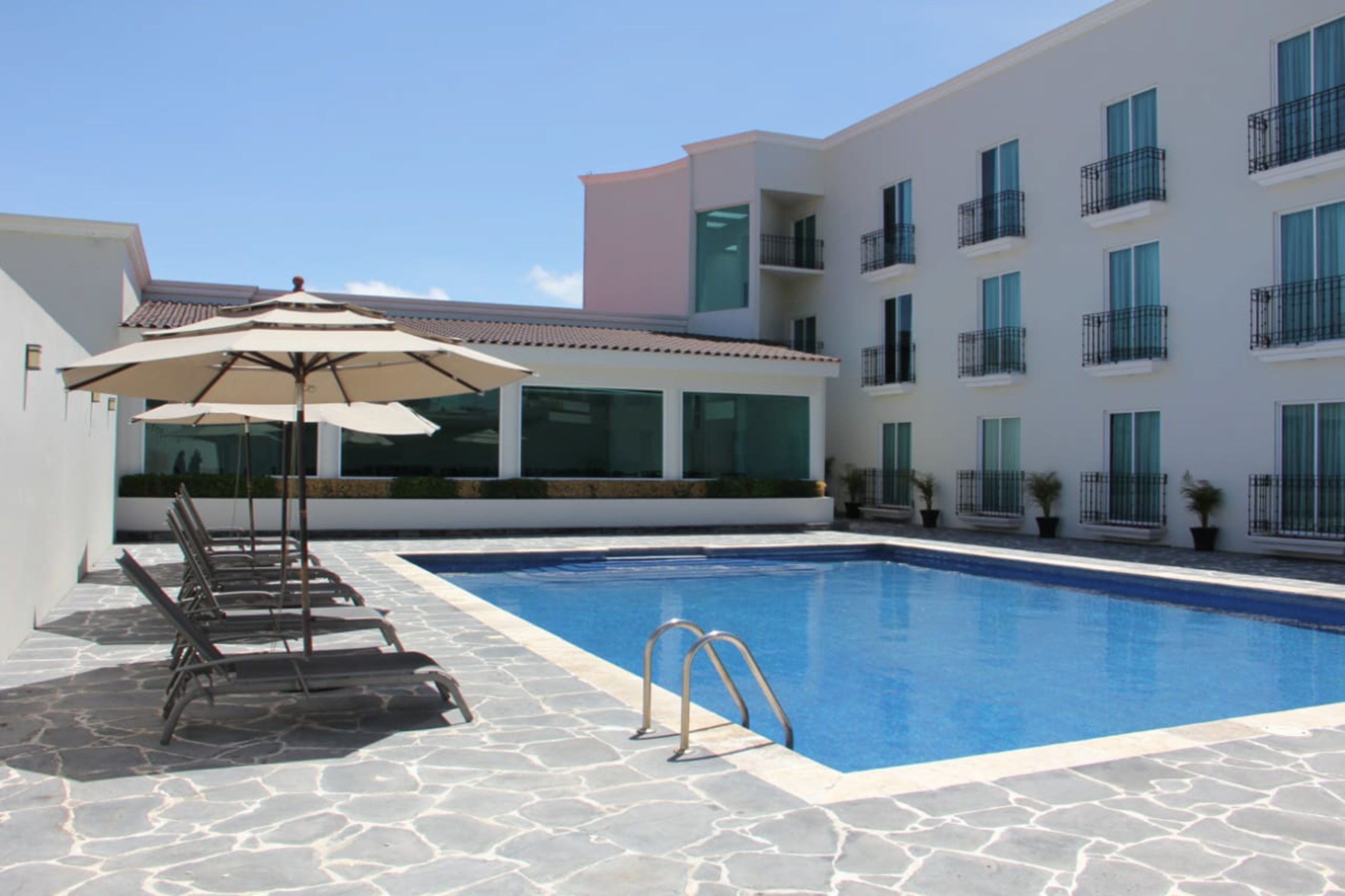 A swimming pool with lounge chairs and umbrellas, surrounded by a building with balconies and large windows.