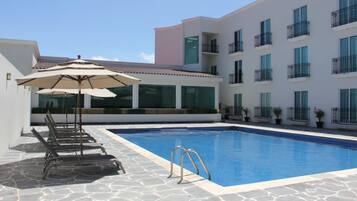 A swimming pool with lounge chairs and umbrellas, surrounded by a building with balconies and large windows.
