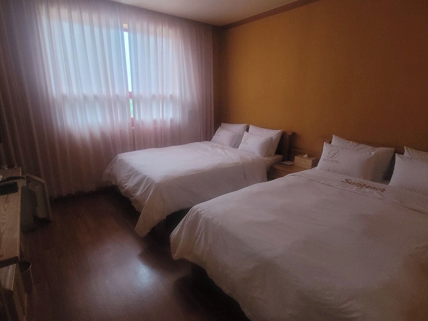 A hotel room with two beds, a wooden floor, and a window with sheer curtains.