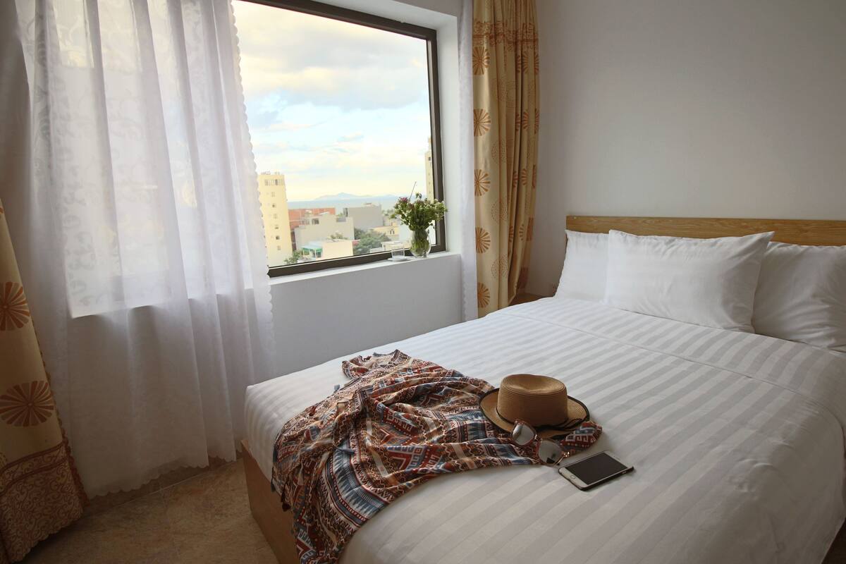 A hotel room with a bed, a window with curtains, a hat, sunglasses, and a smartphone on the bed.