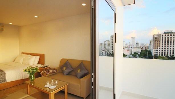Une chambre d’hôtel avec un lit, un canapé, une petite table avec des verres et un vase, et une vue sur la ville à travers une porte vitrée.