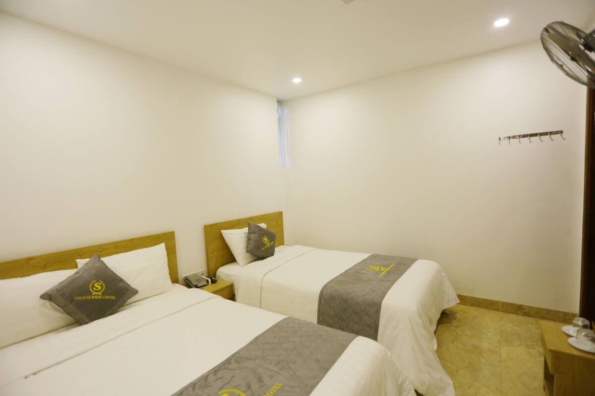 A hotel room with two beds, a wooden headboard, a nightstand, a telephone, a fan, and a coat rack.