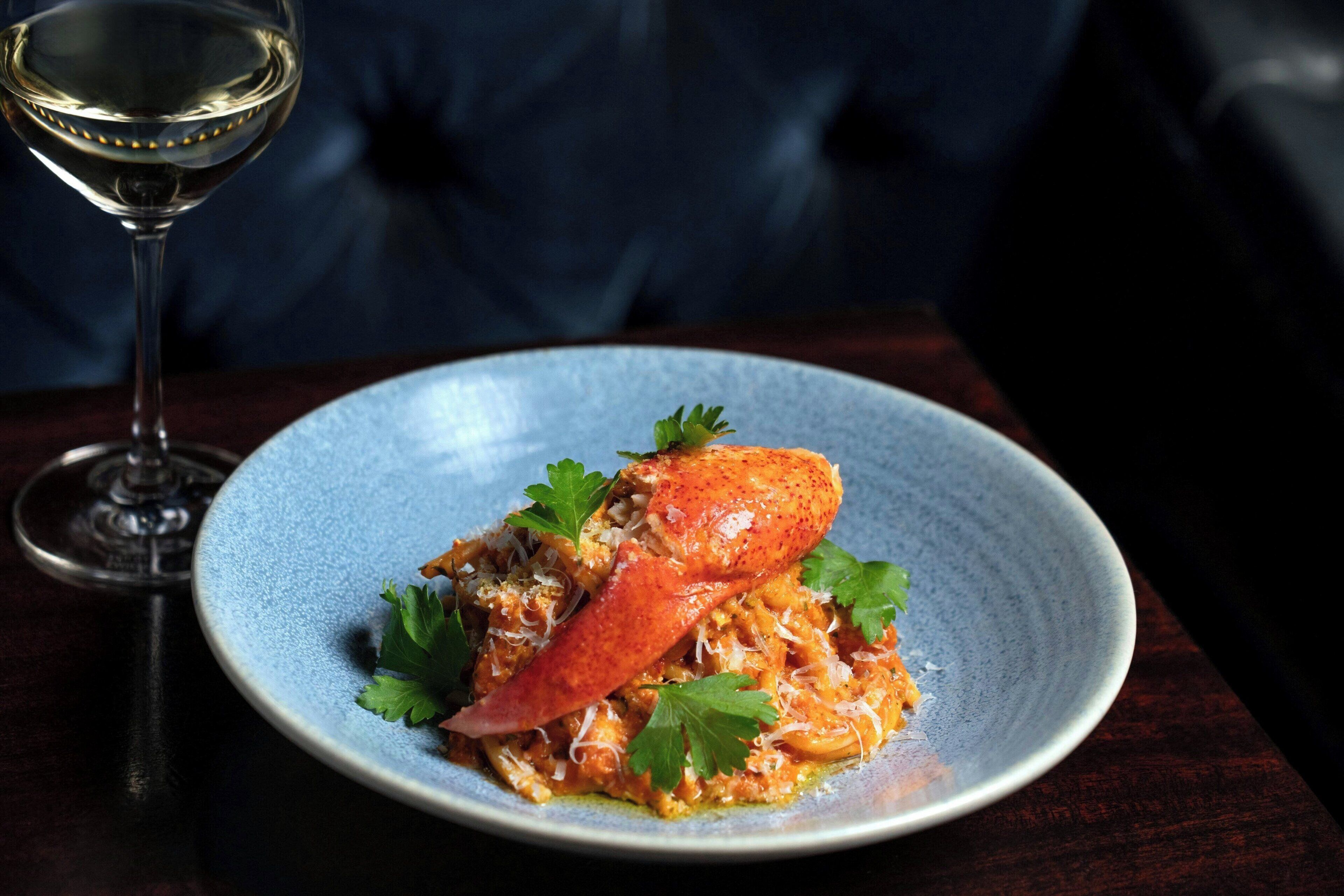A plate of food with a lobster tail and garnishes, accompanied by a glass of white wine on a dark table.