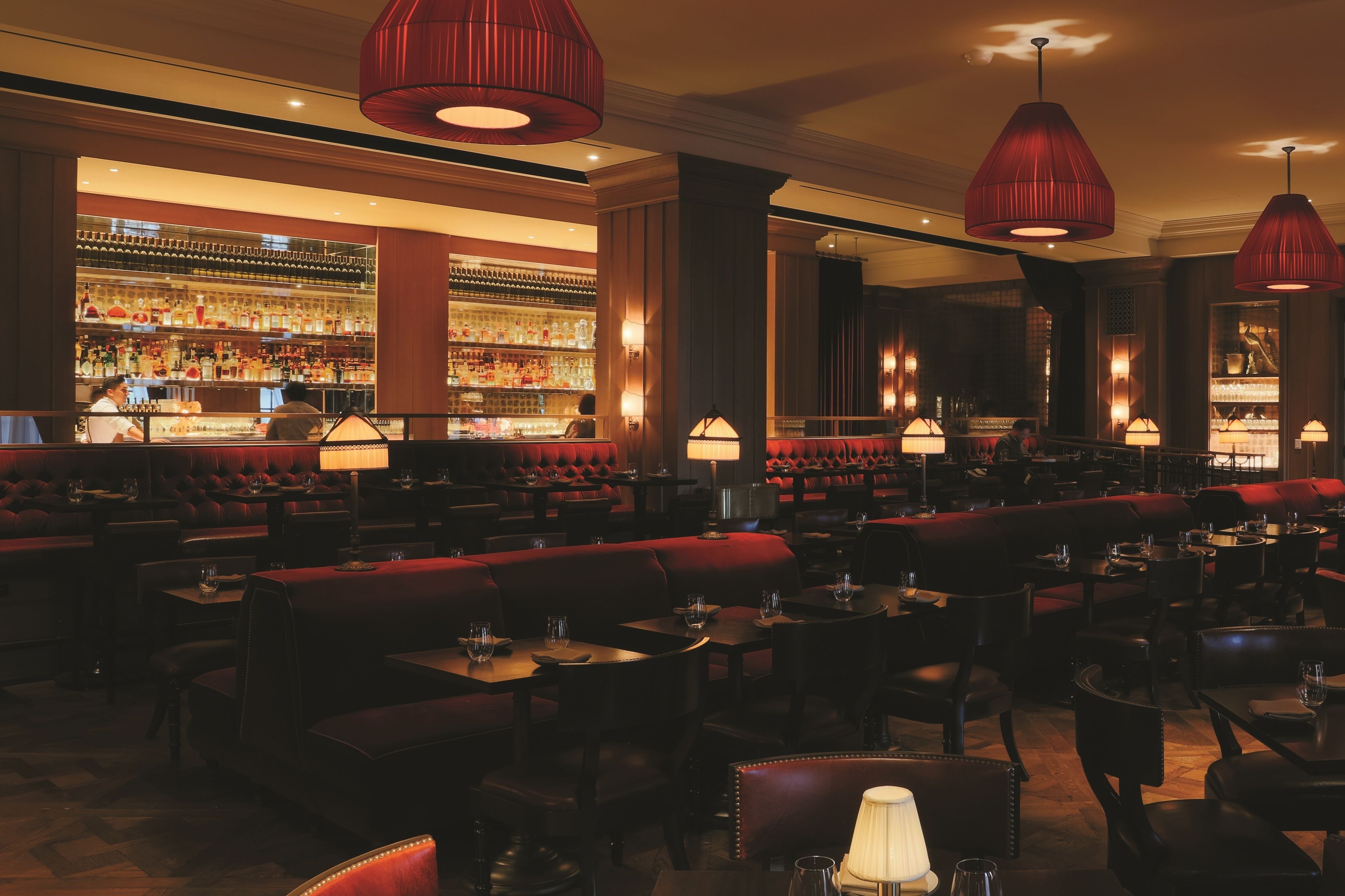 Een ruim restaurant met hoge plafonds, rode gestoffeerde zitmogelijkheden en een bar in de achtergrond.