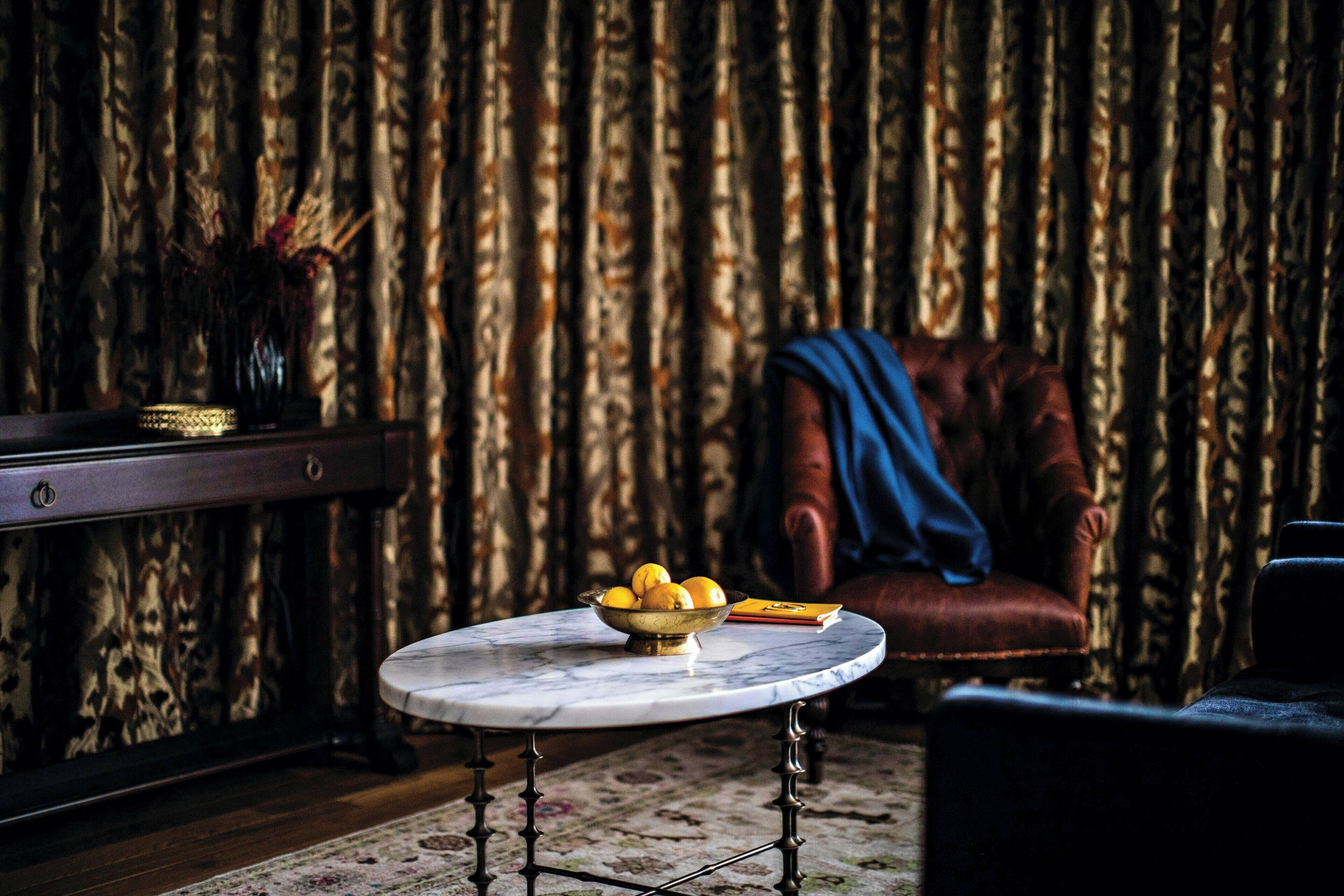 A room with a round table, a bowl of fruit, a leather armchair, and a patterned curtain.