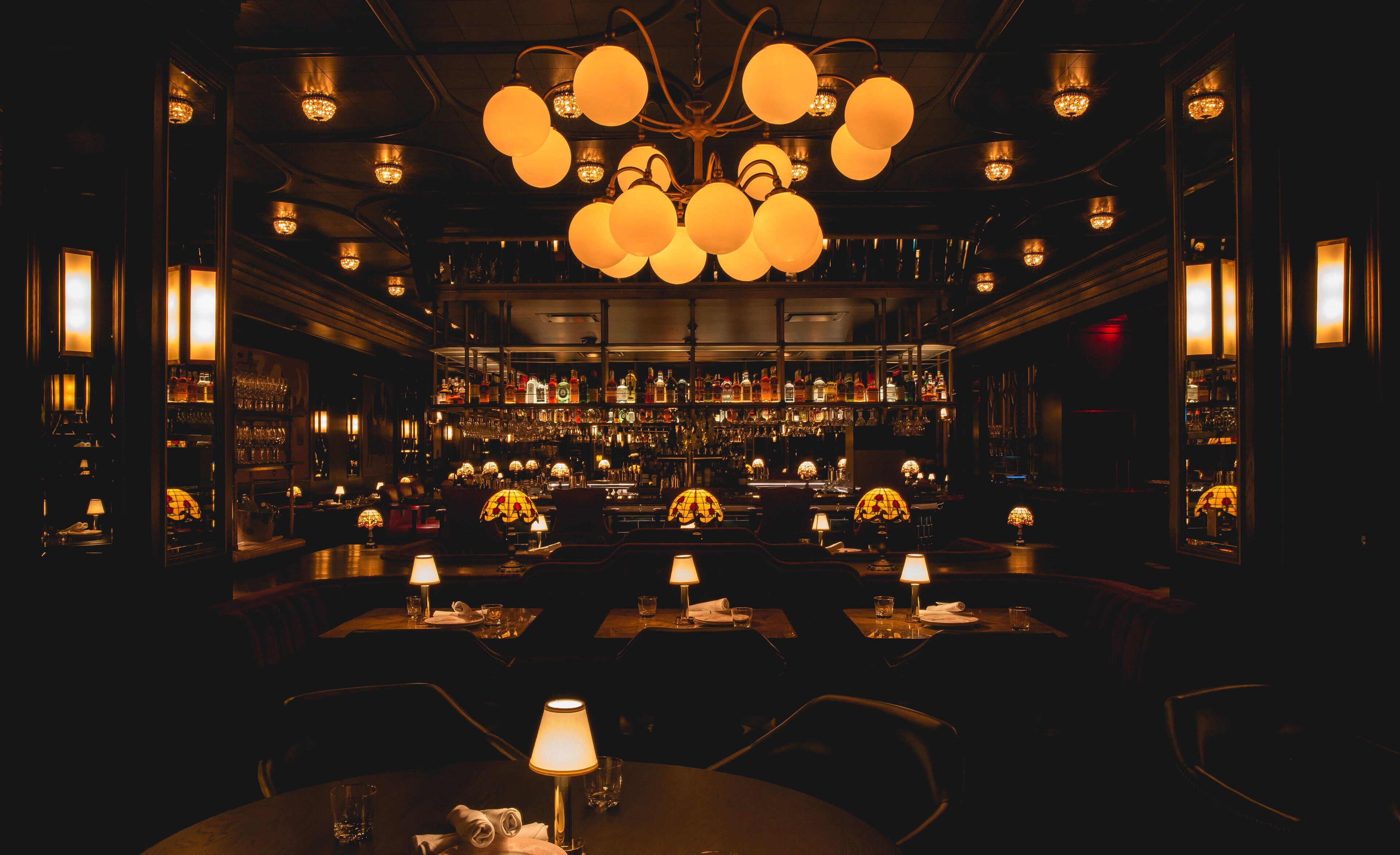 A dimly lit bar with multiple tables, each with a lamp and a bottle. The bar features a variety of bottles and glasses on shelves.