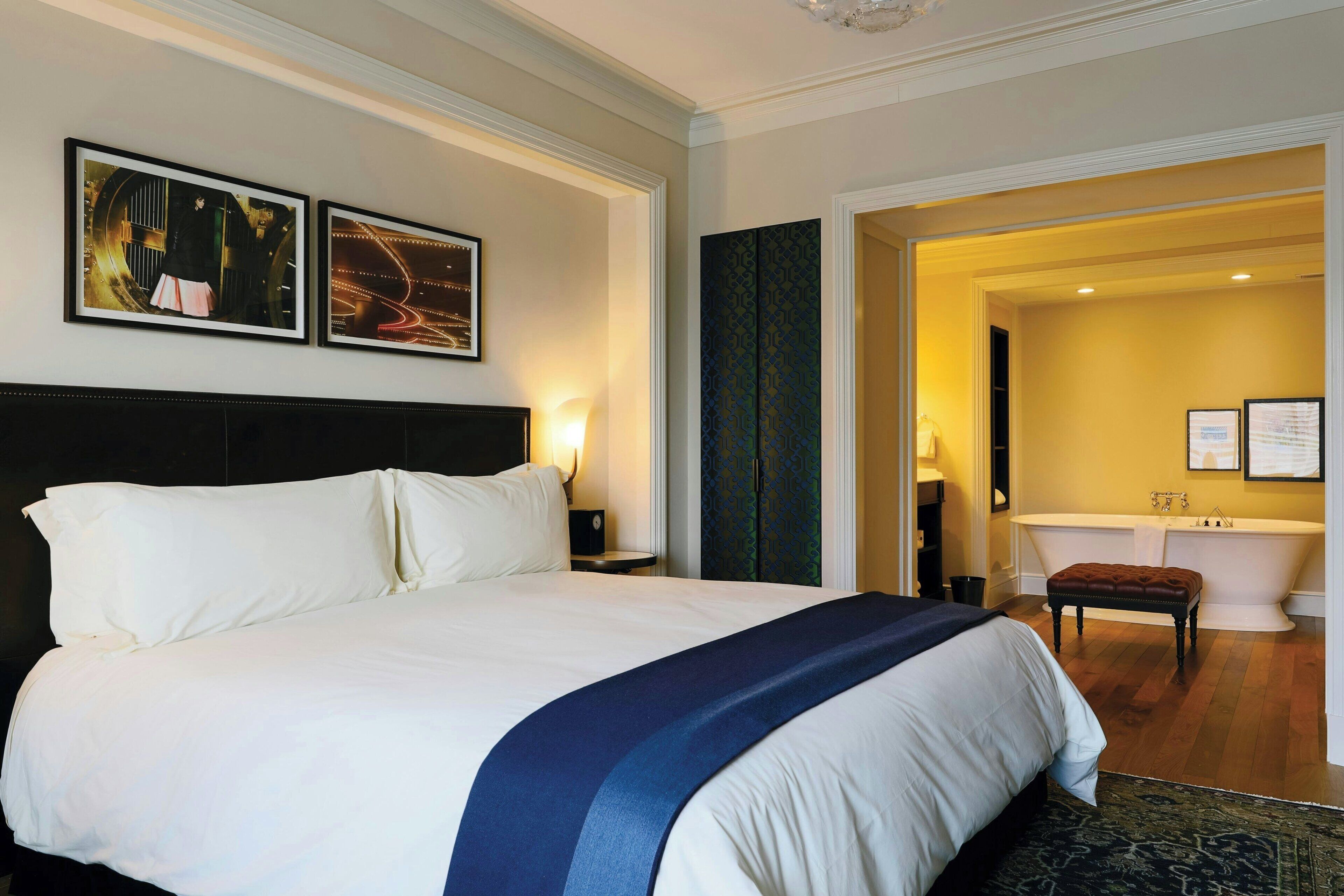A hotel room with a large bed, two wall-mounted artworks, a nightstand, a bench, and a bathroom with a bathtub and a towel rack.